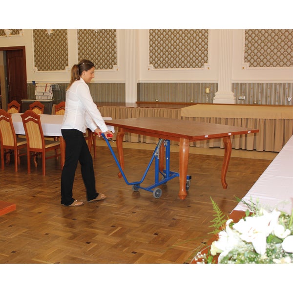 Eine Person bewegt einen großen Holztisch mit einem blauen Tischwagen aus Metall in einem Raum mit Parkettboden.