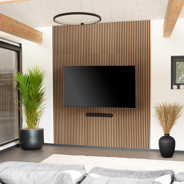Modernes Wohnzimmer mit Wandpaneel aus Holzlamellen hinter einem Fernseher, Deckenbalken aus Holz, Zimmerpflanze und hellem Teppich.