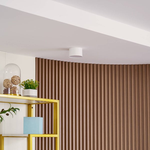 Weißer zylindrischer Deckenaufbaustrahler an einer Decke neben einer Wand mit vertikalen Holzlamellen und einem goldenen Regal.