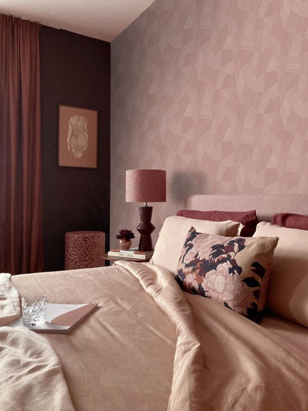 Schlafzimmer mit geometrisch gemusterter Tapete, einem Bett mit Dekokissen, einem Nachttisch mit Lampe und einer dunklen Akzentwand mit Kunstwerk.