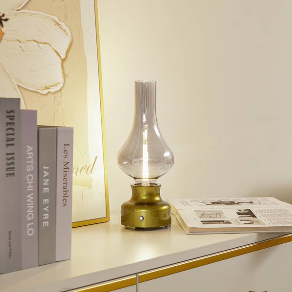 Tragbare LED-Tischleuchte im Vintage-Stil einer Petroleumlampe mit goldenem Sockel und Rauchglas-Schirm auf einem hellen Sideboard mit Büchern.