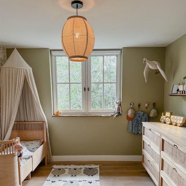 Ein gemütliches Kinderzimmer mit einem Holzbett, einem Himmel, einer Kommode aus Rattan und Holz sowie einer geflochtenen Deckenleuchte.