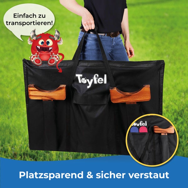 Eine Person trägt eine schwarze Toyfel Transporttasche für ein Holzspiel mit Tragegriffen und Außentaschen für Spielzubehör auf einer Wiese.