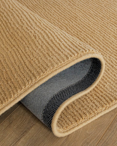 Senffarbener gerippter Teppich mit umgeschlagener Ecke auf Holzboden, zeigt die strukturierte Oberfläche und die dunkle Unterseite.