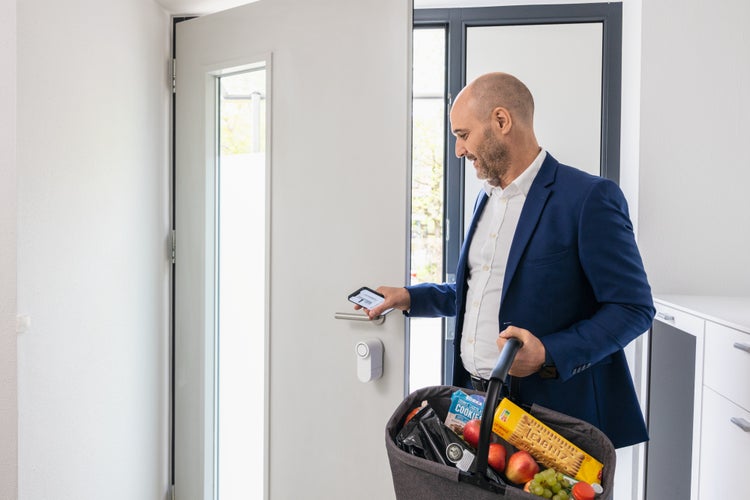 Ein Mann öffnet eine weiße Haustür mit einem Smartphone und einem elektronischen Türschloss, während er einen Einkaufskorb trägt.