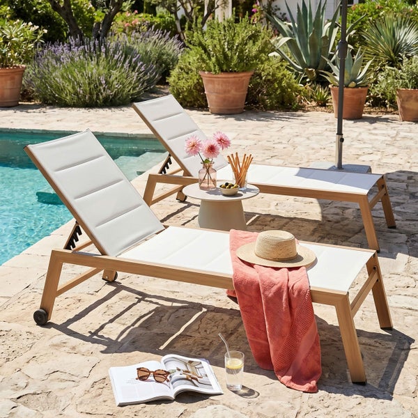 Zwei Sonnenliegen aus Holz mit Polstern auf einer Steinterrasse neben einem Swimmingpool in einem Garten.