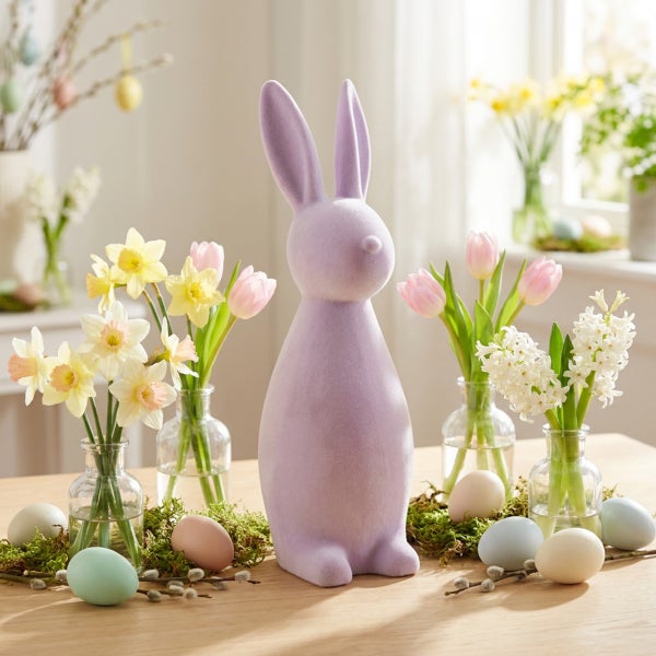 Osterdekoration mit einem flauschigen Osterhasen auf einem Holztisch, umgeben von Blumen in Glasvasen und dekorativen Ostereiern.
