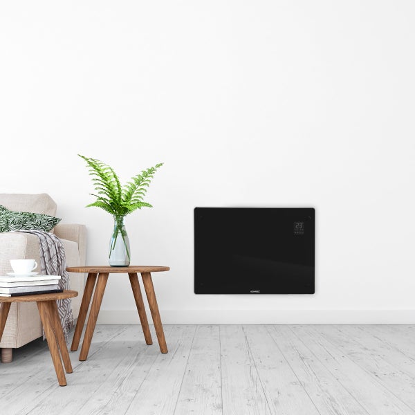 Moderne Glasheizung mit Digitalanzeige und Vonroc Logo an einer Wohnzimmerwand neben einem Couchtisch mit Pflanze und Sofa.
