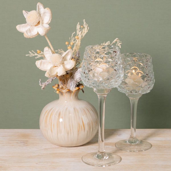 Dekorative Szene mit zwei hohen Teelichthaltern aus strukturiertem Glas und einer runden Keramikvase mit Trockenblumen auf einer Holzoberfläche.