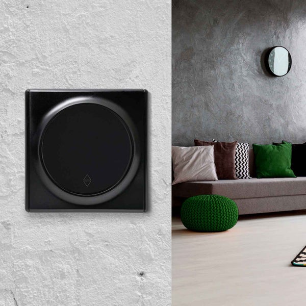 Quadratischer Rollladenschalter mit rundem Bedienteil montiert an einer hellen Wand neben einem modern eingerichteten Wohnzimmer.