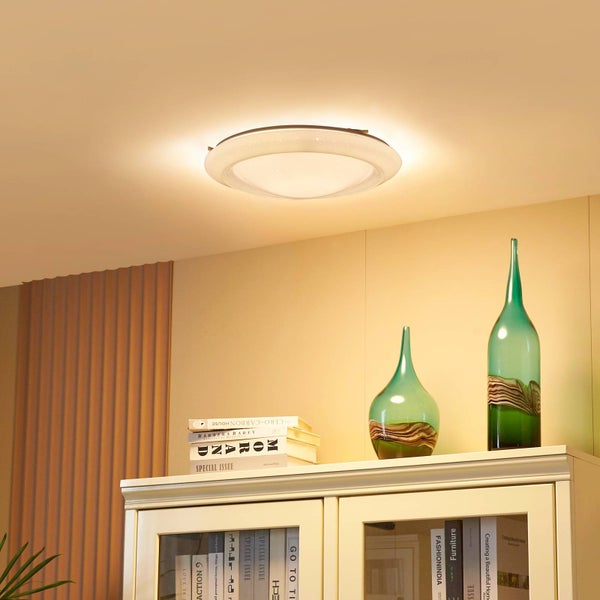 Moderne runde LED-Deckenleuchte mit Sternenhimmel-Effekt und warmweißem Licht in einem hellen Wohnraum über einem Schrank mit Deko-Glasvasen.