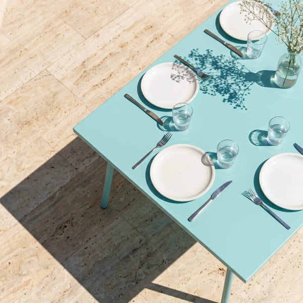 Gedeckter hellblauer Gartentisch auf einer Terrasse mit Natursteinfliesen, ausgestattet mit Tellern, Gläsern und Besteck für vier Personen.