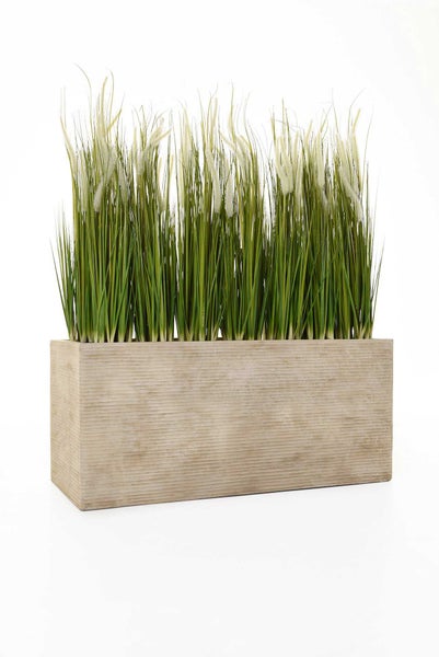 Künstliches Gras mit weißen Blütenwedeln in einem rechteckigen Pflanzkübel mit gerippter Struktur.