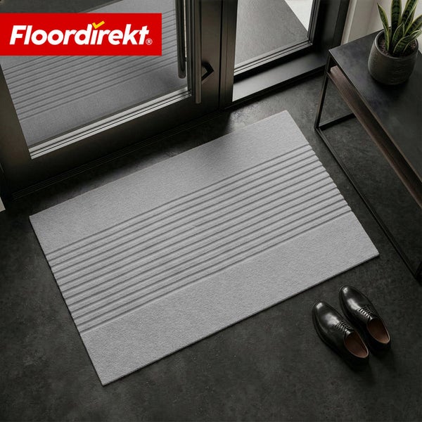 Floordirekt Logo. Schmutzfangmatte vor einer Glastür mit Rillenmuster, Schuhen und Zimmerpflanze in einem modernen, hellen Eingangsbereich.