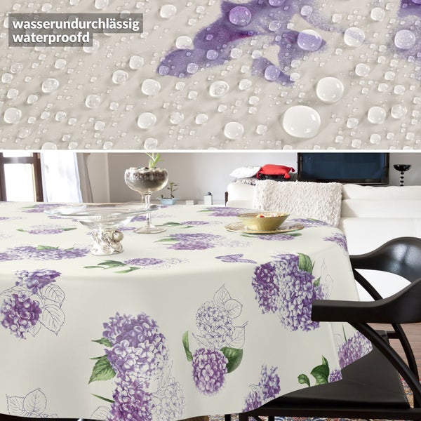 Runde Tischdecke mit lila Hortensien-Muster in hellem Wohnzimmer. Detailansicht zeigt wasserundurchlässiges Material mit Wassertropfen.