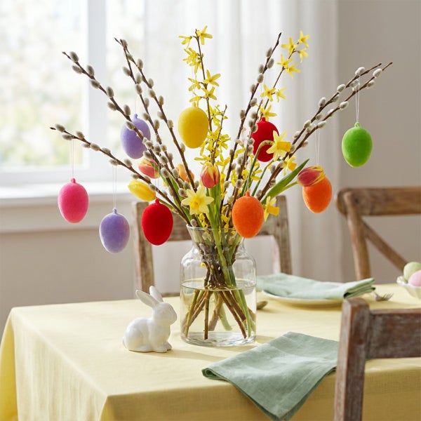 Osterstrauß mit Weidenkätzchen und Forsythien in einer Glasvase, dekoriert mit Ostereiern und einer weißen Osterhasenfigur auf einem Tisch.