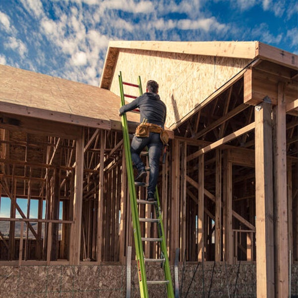 Handwerker mit Werkzeuggürtel auf einer Anlegeleiter am Holzrahmen eines im Bau befindlichen Hauses.