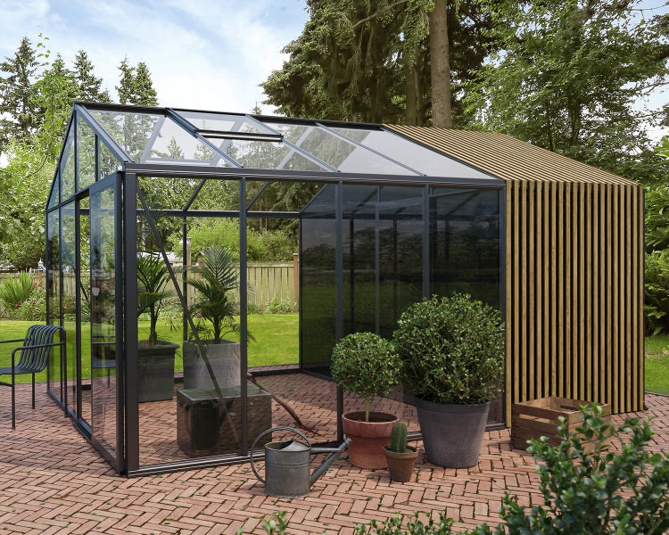 Modernes Gewächshaus mit schwarzem Metallrahmen und Glaselementen sowie einem Anbau aus Holzlamellen auf einer Ziegelterrasse im Garten.