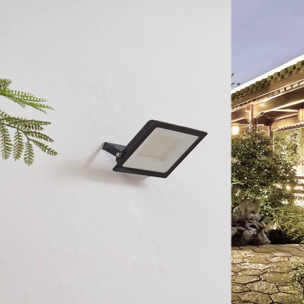 Schwarzer LED-Außenstrahler an einer weißen Wand montiert, im Hintergrund ein Garten mit Steinweg und Pflanzen.