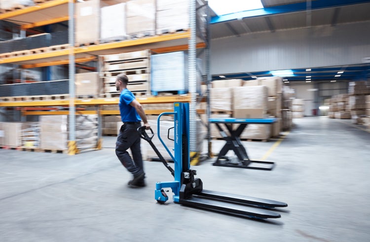 Person bewegt einen blauen manuellen Hochhubwagen in einem hellen Lager mit Industrieregalen und palettierten Waren.