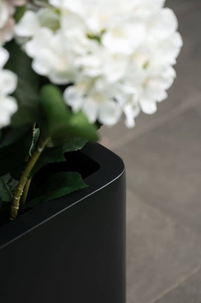 Nahaufnahme eines mattschwarzen Pflanzgefäßes mit abgerundeten Ecken, bestückt mit künstlichen weißen Blumen und grünen Blättern.