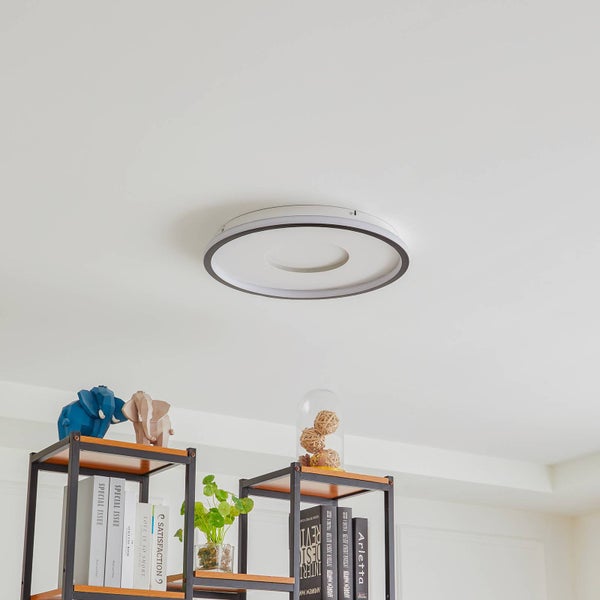Runde Deckenleuchte mit dunklem Rand an einer weißen Zimmerdecke über Regalen mit Dekorationsartikeln und Büchern.