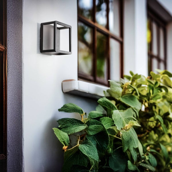 Moderne würfelförmige Außenwandleuchte in Schwarz an einer Hauswand neben einem Fenster und grünen Pflanzen.
