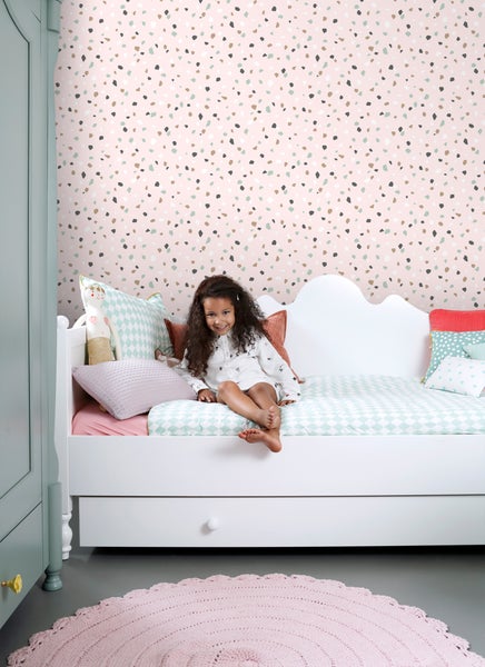 Mädchen sitzt in einem hellen Kinderzimmer mit gemusterter Tapete und weißem Bett