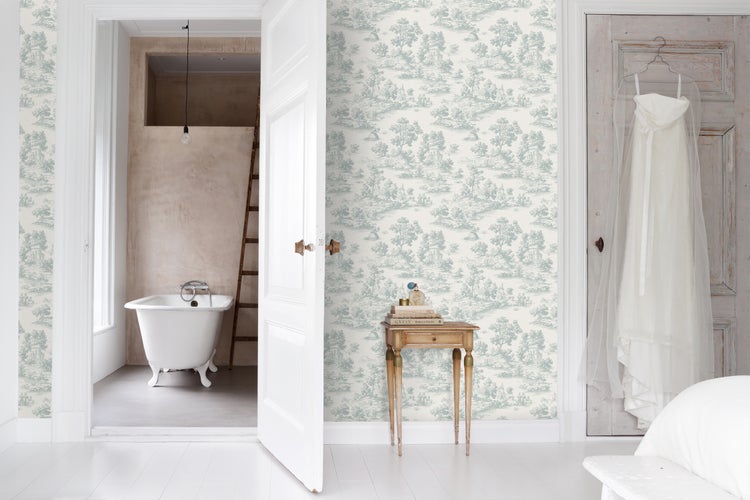 Zimmer mit Toile-de-Jouy-Tapete mit Landschaftsmuster, Beistelltisch aus Holz und Blick ins Bad mit freistehender Badewanne.