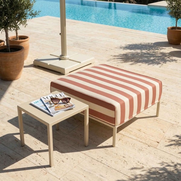 Ein gepolsterter Gartenhocker mit gestreifter Auflage und ein Beistelltisch auf einer Terrasse am Pool mit Olivenbäumen und Sonnenschirmfuß.