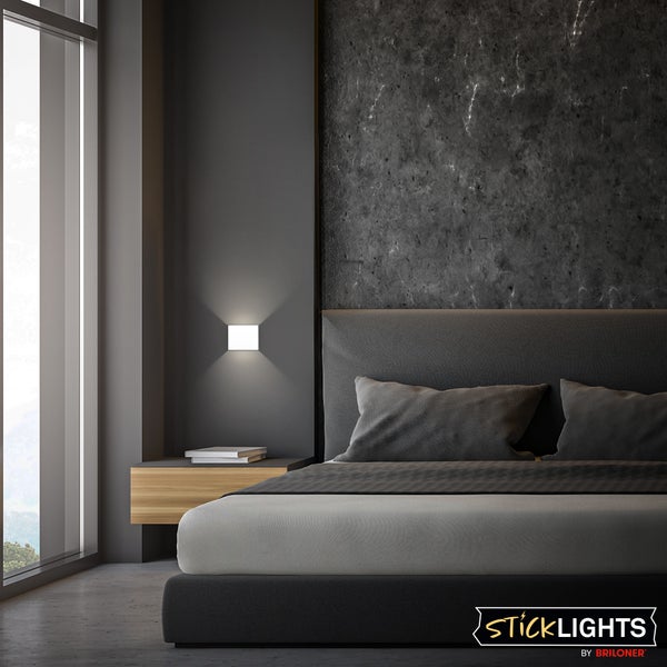 Quadratische weiße Wandleuchte in einem modernen Schlafzimmer mit grauem Bett und Wand in Steinoptik. StickLights Logo.