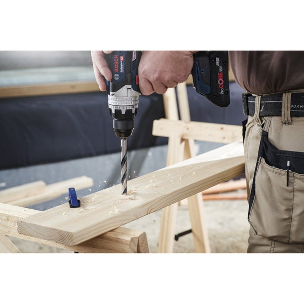 Ein Mann bohrt mit einem Akku-Bohrschrauber ein Loch in ein Holzbrett. Im Hintergrund steht ein Holzbock.