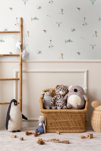 Helle Kindertapete mit Zirkusmotiven in einem Kinderzimmer mit einer Holzleiter, einem geflochtenen Korb und diversen Stofftieren auf dem Teppich.