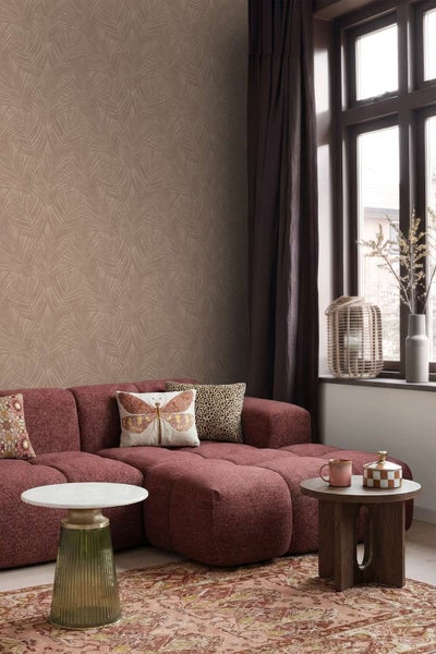 Wohnzimmer mit Vliestapete mit Palmenblattmuster, rotes Modulsofa, Beistelltische, Teppich und Deko, einladende und moderne Raumatmosphäre.