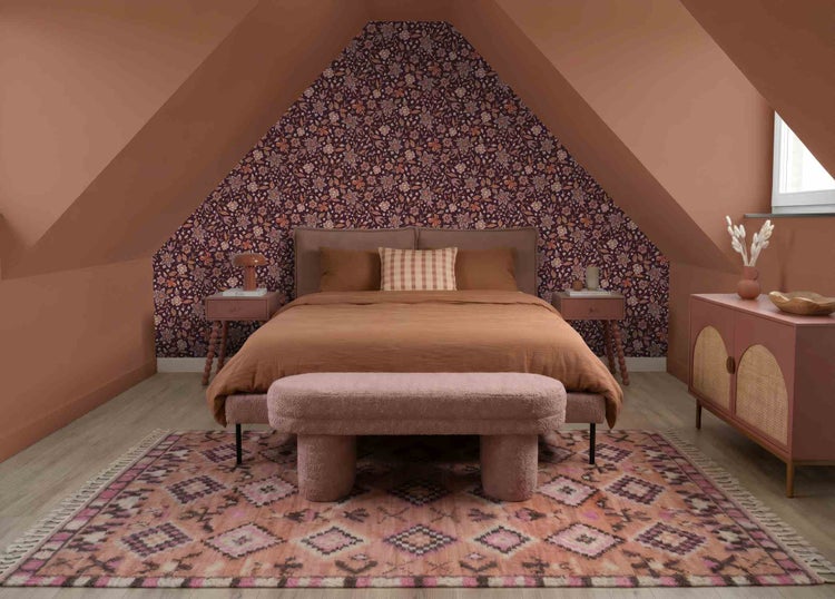 Schlafzimmer unter Dachschräge mit floraler Tapete, terracottafarbener Wandfarbe, Doppelbett, gemustertem Teppich und Polsterbank.