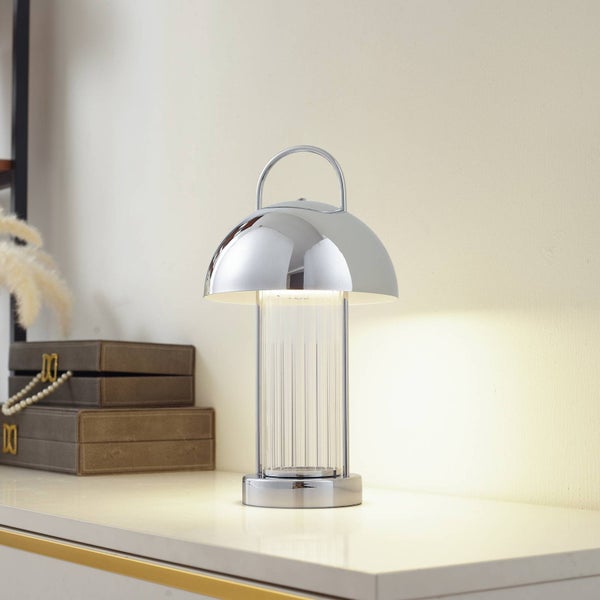 Moderne Tischleuchte in Chrom-Optik mit kuppelförmigem Schirm und Tragegriff auf einem hellen Sideboard neben dekorativen Boxen.