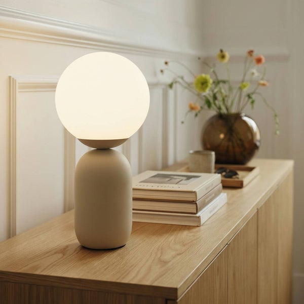 Moderne Tischleuchte mit kugelförmigem weißem Schirm und beigem Sockel auf einer hellen Holzkommode in einem hellen, organisierten Raum.