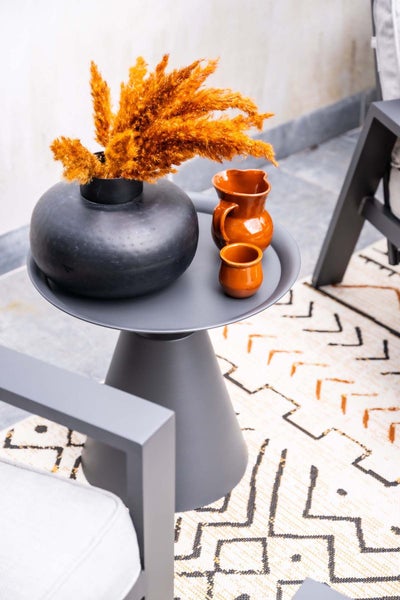 Moderner Beistelltisch aus Metall mit kegelförmigem Fuß, dekoriert mit einer Vase, Trockengras und Keramik auf einem gemusterten Teppich.
