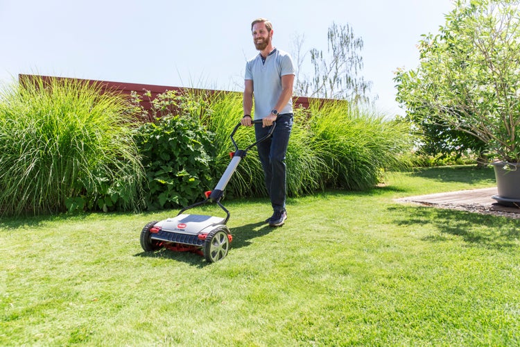 Ein Mann schiebt einen handbetriebenen Spindelmäher über eine Rasenfläche in einem sonnigen Garten mit grünen Pflanzen und Sträuchern.
