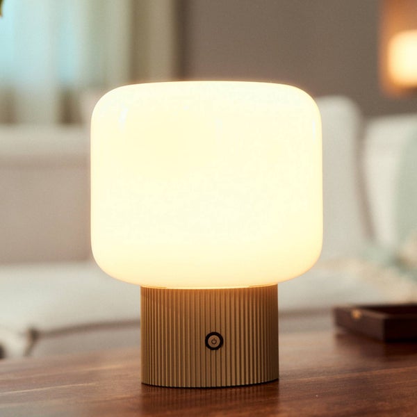 Moderne Tischleuchte mit geripptem, goldfarbenem Sockel und abgerundetem, weißem Schirm auf einem Holztisch in einem hellen Raum.