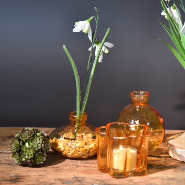 Frühlingshafte Tischdekoration mit bernsteinfarbenen Glasvasen, Schneeglöckchen und einem Teelichthalter auf Holzoberfläche.