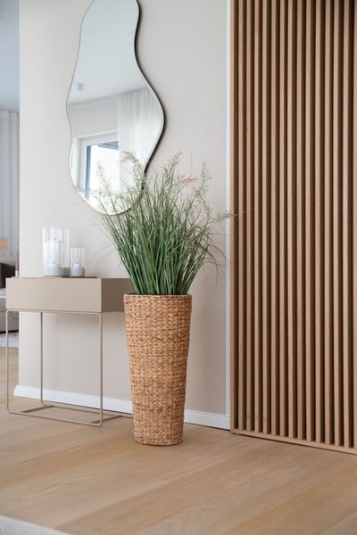 Modernes Interieur mit hoher Kunstpflanze im geflochtenen Topf, Konsolentisch, Wandspiegel und Wandpaneel aus Holzlamellen auf Holzboden.
