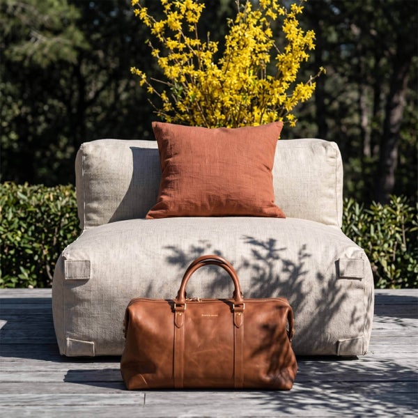 Beige Loungesessel für den Außenbereich auf einer Holzterrasse mit einem orangefarbenen Kissen und einer braunen Ledertasche davor.