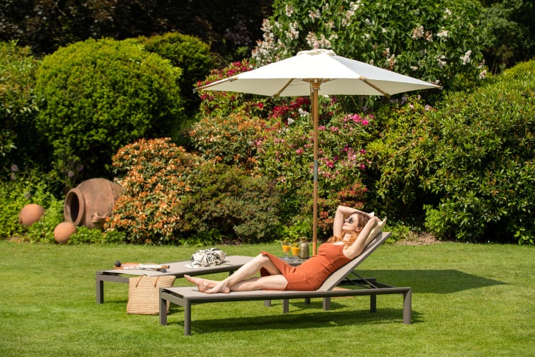 Garten mit zwei Sonnenliegen und einem Sonnenschirm auf einem Rasen, Frau entspannt in einer Sonnenliege, sonnige Gartenatmosphäre.