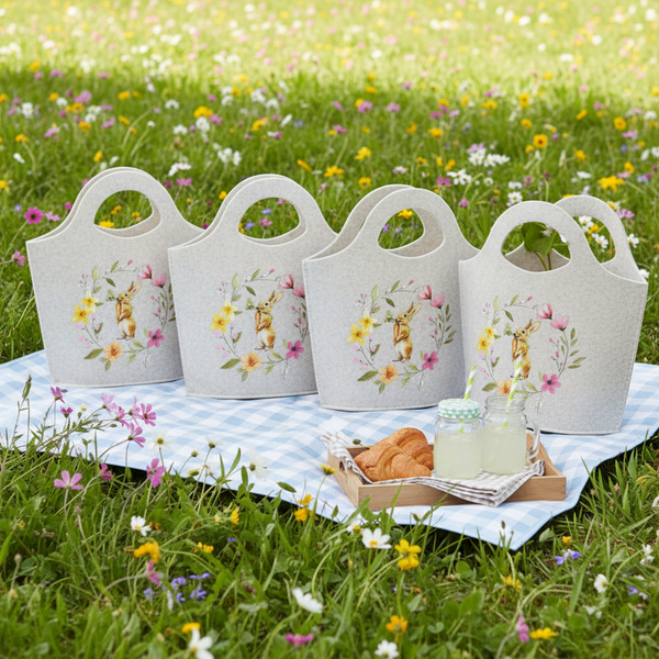 Vier Filztaschen mit Osterhasen-Motiv auf einer Picknickdecke in einer Blumenwiese, dazu Croissants und Getränke auf einem Holztablett.