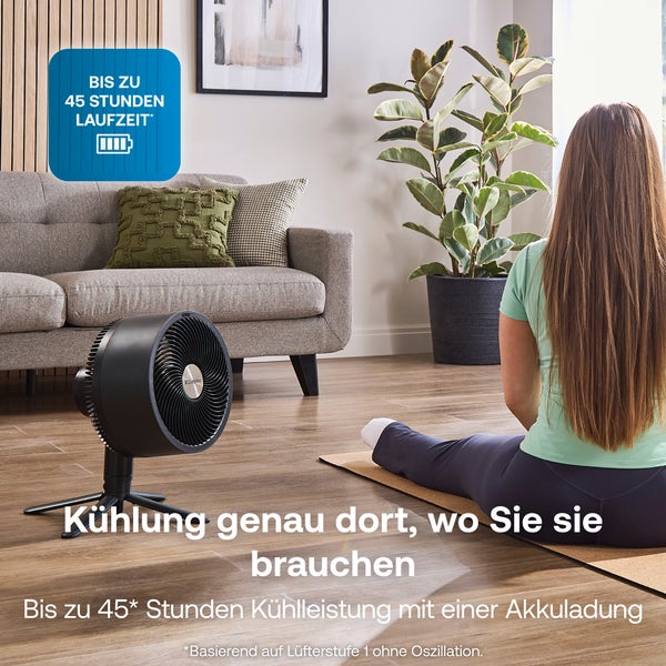 Akkubetriebener schwarzer Dimplex Bodenventilator in hellem Wohnzimmer neben einer Frau auf einer Yogamatte, Dimplex Logo, bis zu 45 Stunden Laufzeit.