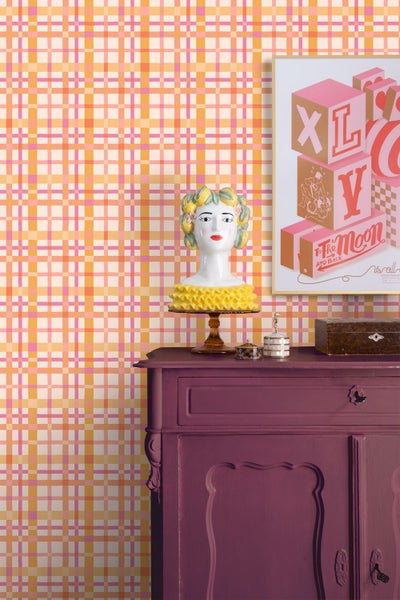 Vliestapete mit gelb-rosa Karomuster in einem Wohnzimmer über einer lila Kommode mit dekorativer Büste und gerahmtem Bild.