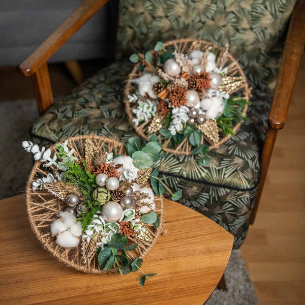 Dekorative Gestecke mit Baumwolle, Tannenzapfen und Kugeln auf einem Tisch und einem Stuhl.