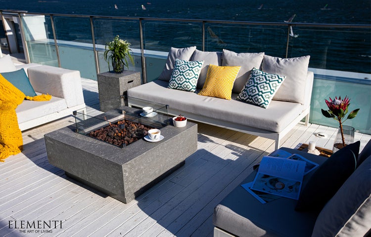 Moderner Gaskamin-Tisch aus Beton mit Glasschutz auf einer hellen Terrasse mit organisierten Gartenmöbeln und Blick auf das Meer.
