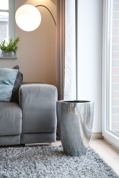 Modernes Wohnzimmer mit einem hohen, silbernen Pflanzkübel aus Metall auf einem grauen Teppich neben einem Sofa und einer Bogenlampe.